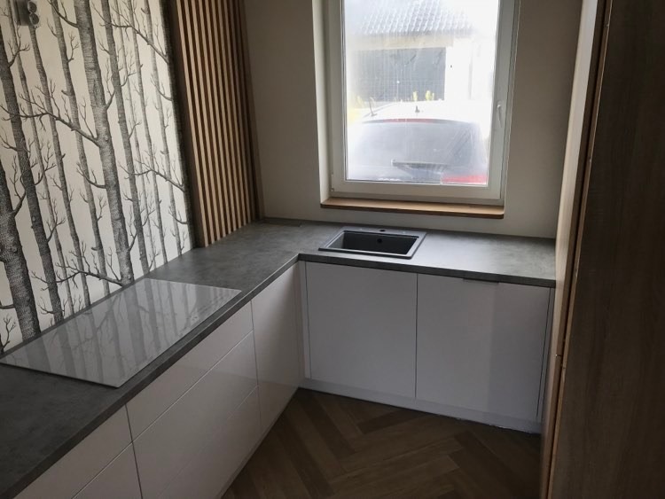 Nowoczesna kuchnia z białymi szafkami, szarym blatem i tapetą z motywem drzew, uzupełniona o drewniane lamele ścienne i parkiet w jodełkę.