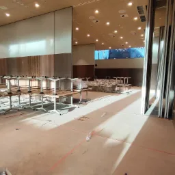 Przestronne wnętrze sali konferencyjnej w trakcie przygotowań, widoczne stoły z białymi nogami i drewnianymi blatami, rozłożone po całej powierzchni, z widocznymi elementami instalacji pod sufitem