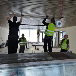 GRUPA HOFMAN - INSTALACJE - Pracownicy w odblaskowych kamizelkach montują drewniane panele sufitowe w nowoczesnym wnętrzu, widoczne przewody elektryczne i biała puszka instalacyjna.