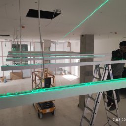 GRUPA HOFMAN - INSTALACJE - Montera na drabinie poziomuje laserowo podwieszane, długie panele oświetleniowe LED w surowym wnętrzu biurowym. Widoczny podnośnik i zielone linie lasera.