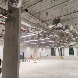 GRUPA HOFMAN - INSTALACJE - Wnętrze hali w trakcie prac wykończeniowych. Widoczne instalacje pod sufitem, kolumna w szpachli, drabina i pracownik w oddali. Oświetlenie ledowe zamontowane.