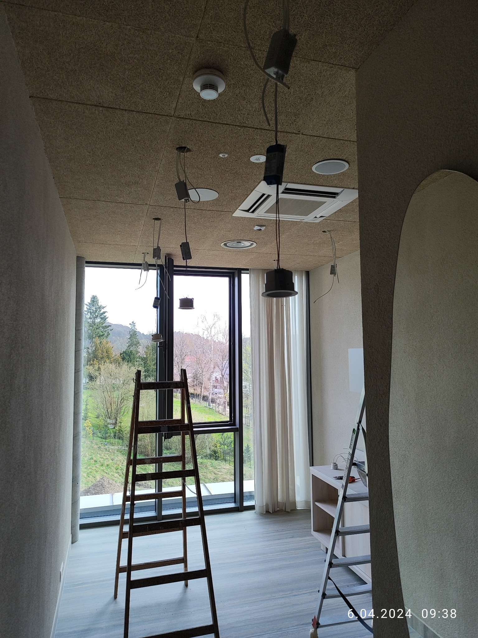 Wnętrze w trakcie prac w nowym budownictwie we Wrocławiu. Widoczne drabiny, okno z widokiem na zieleń, sufit z niedokończonymi punktami oświetleniowymi i przewodami.