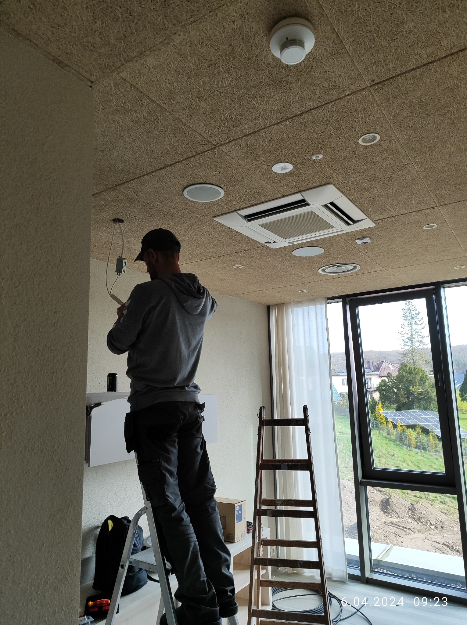 Elektryk na drabinie podłącza przewody w nowym budynku, widoczny sufit z oświetleniem i klimatyzacją, za oknem panele słoneczne.