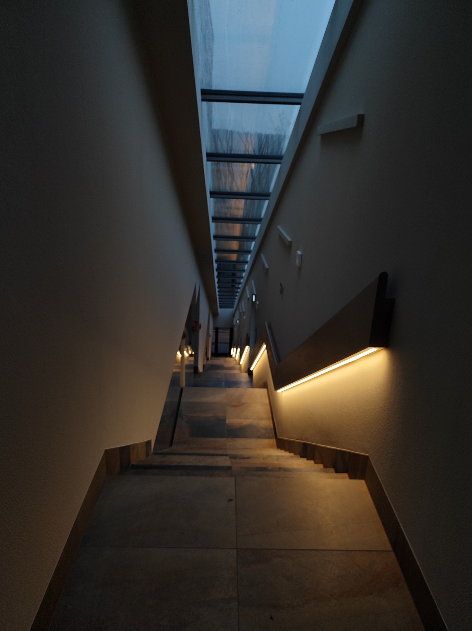 Długi korytarz ze schodami w dół, oświetlony liniowymi lampami LED zamontowanymi na ścianach i naturalnym światłem z przeszklonego dachu.