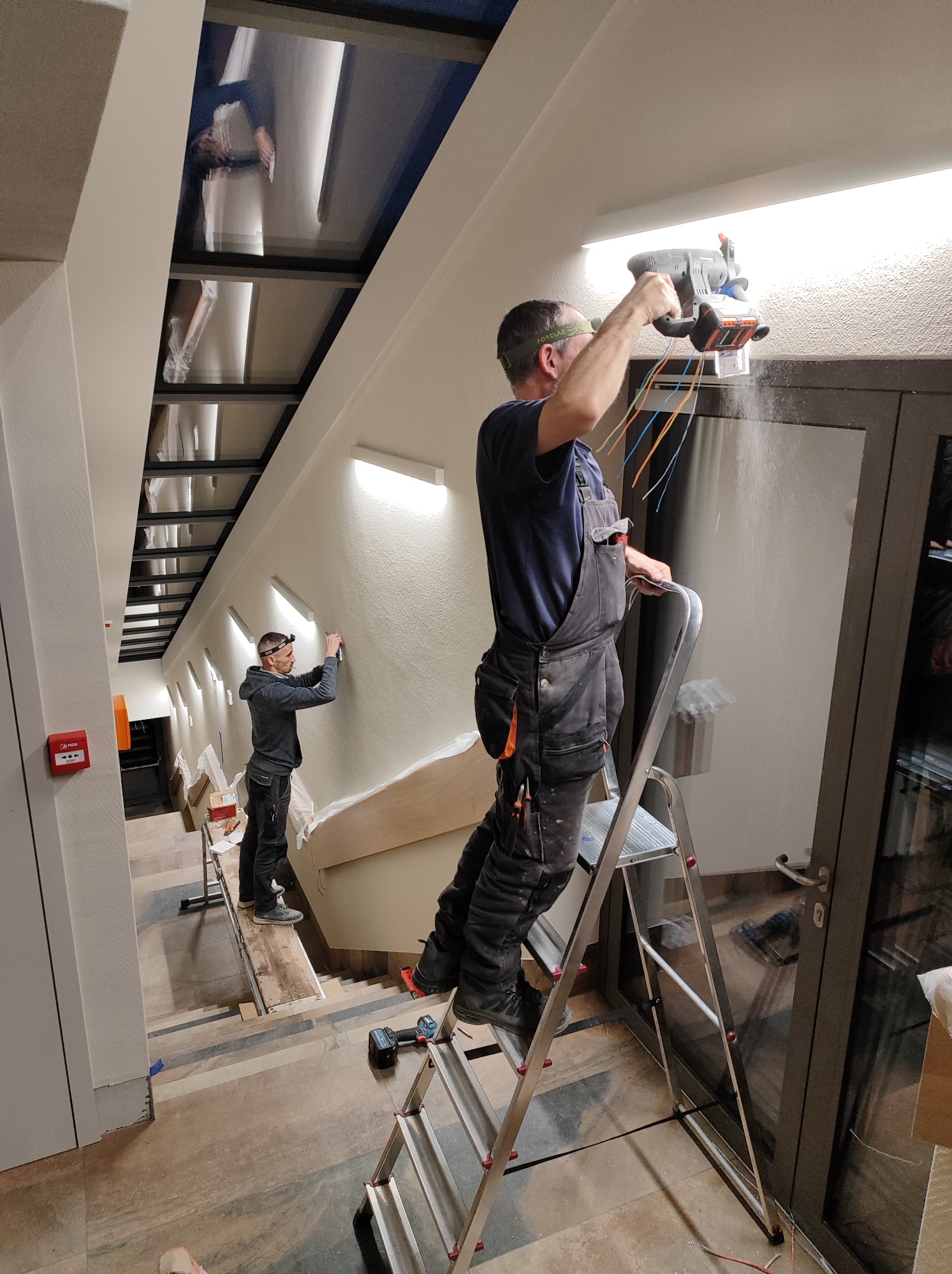 Elektryk na drabinie tnie otwór w ścianie pod oświetlenie, widoczne kable i pył, drugi elektryk montuje lampę w tle na schodach.