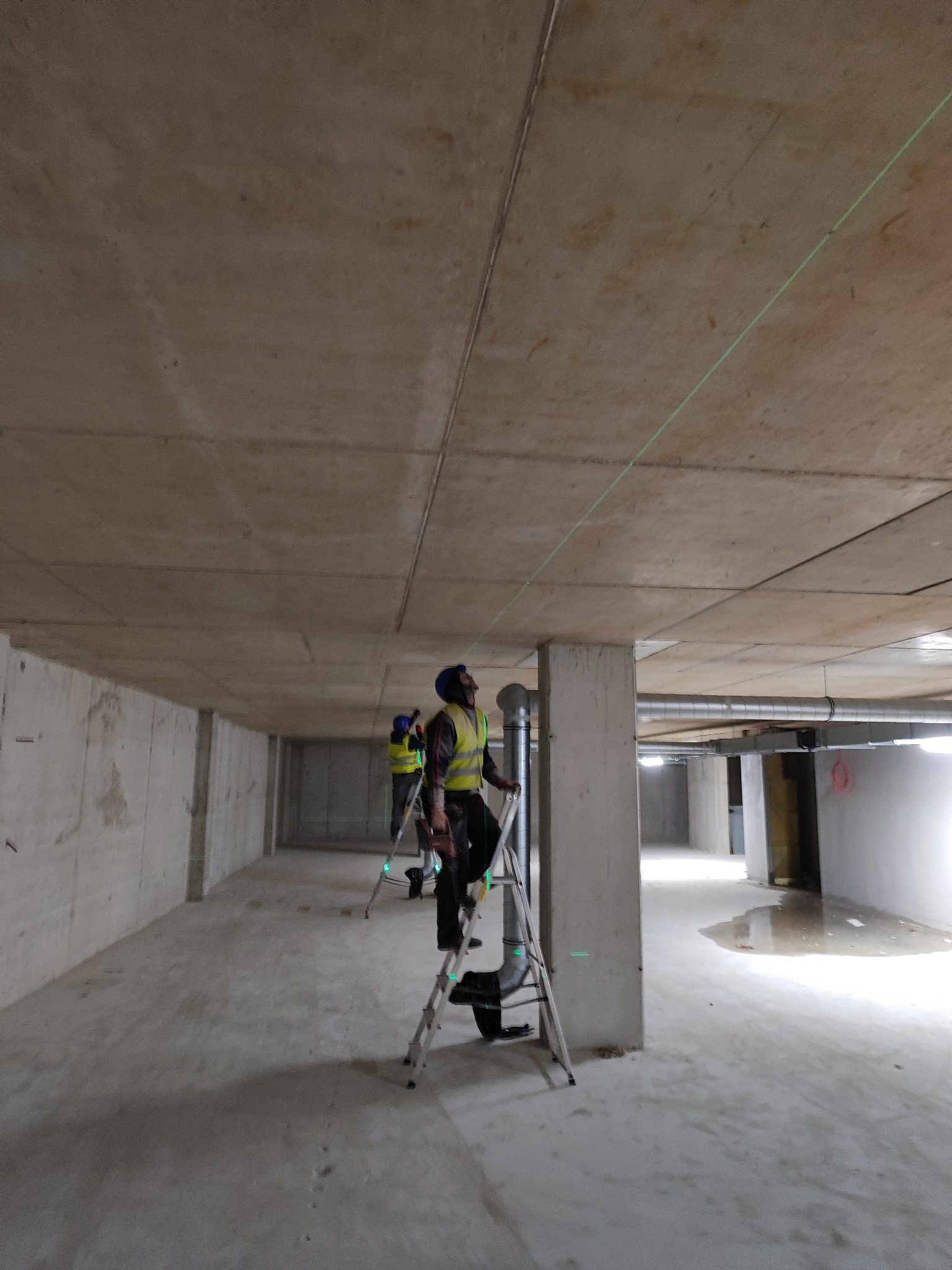 Dwóch pracowników na drabinach ustawia laserowy poziom na surowym betonowym suficie w pustym, niedokończonym pomieszczeniu z widocznymi rurami wentylacyjnymi.