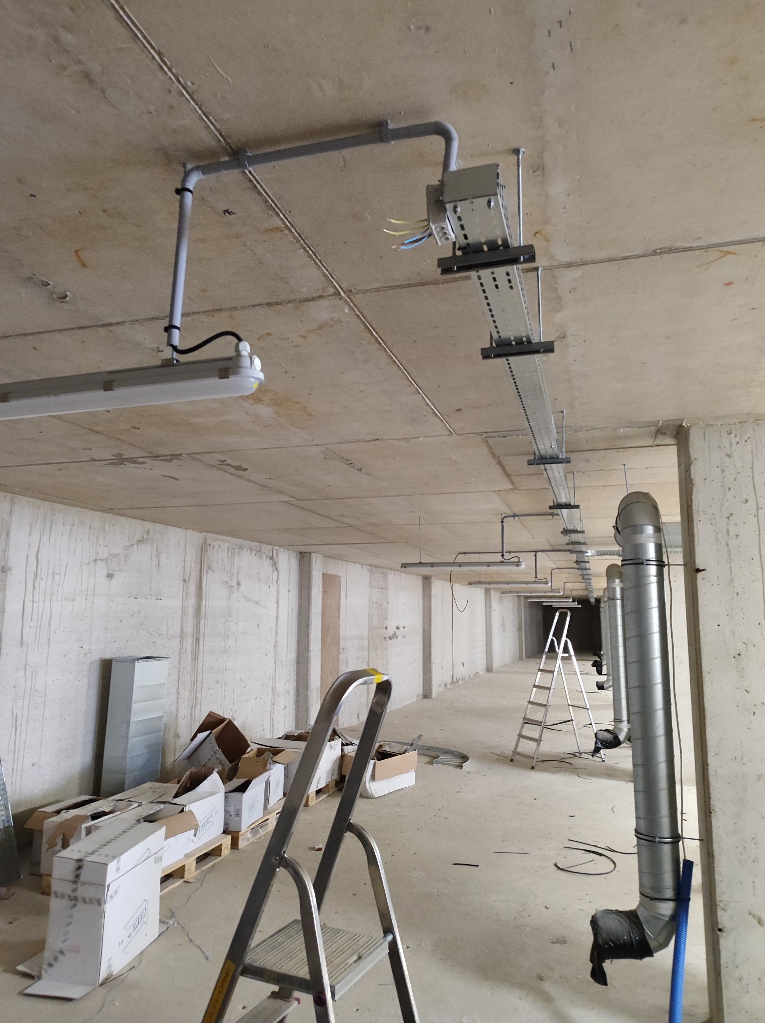 Wnętrze surowego budynku w trakcie prac instalacyjnych: widoczne rury wentylacyjne, koryta kablowe pod sufitem, oświetlenie fluorescencyjne i rozłożone kartony na paletach, drabina aluminiowa...