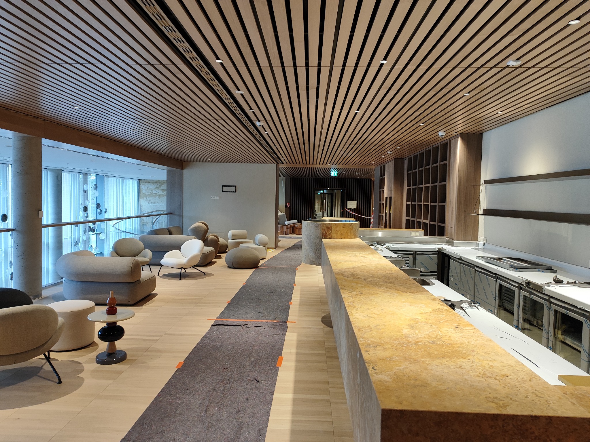 Wnętrze w trakcie wykańczania: widoczny drewniany sufit z oświetleniem punktowym, meble wypoczynkowe w tle oraz fragment baru z urządzeniami chłodniczymi w trakcie montażu, podłoga zabezpieczona...