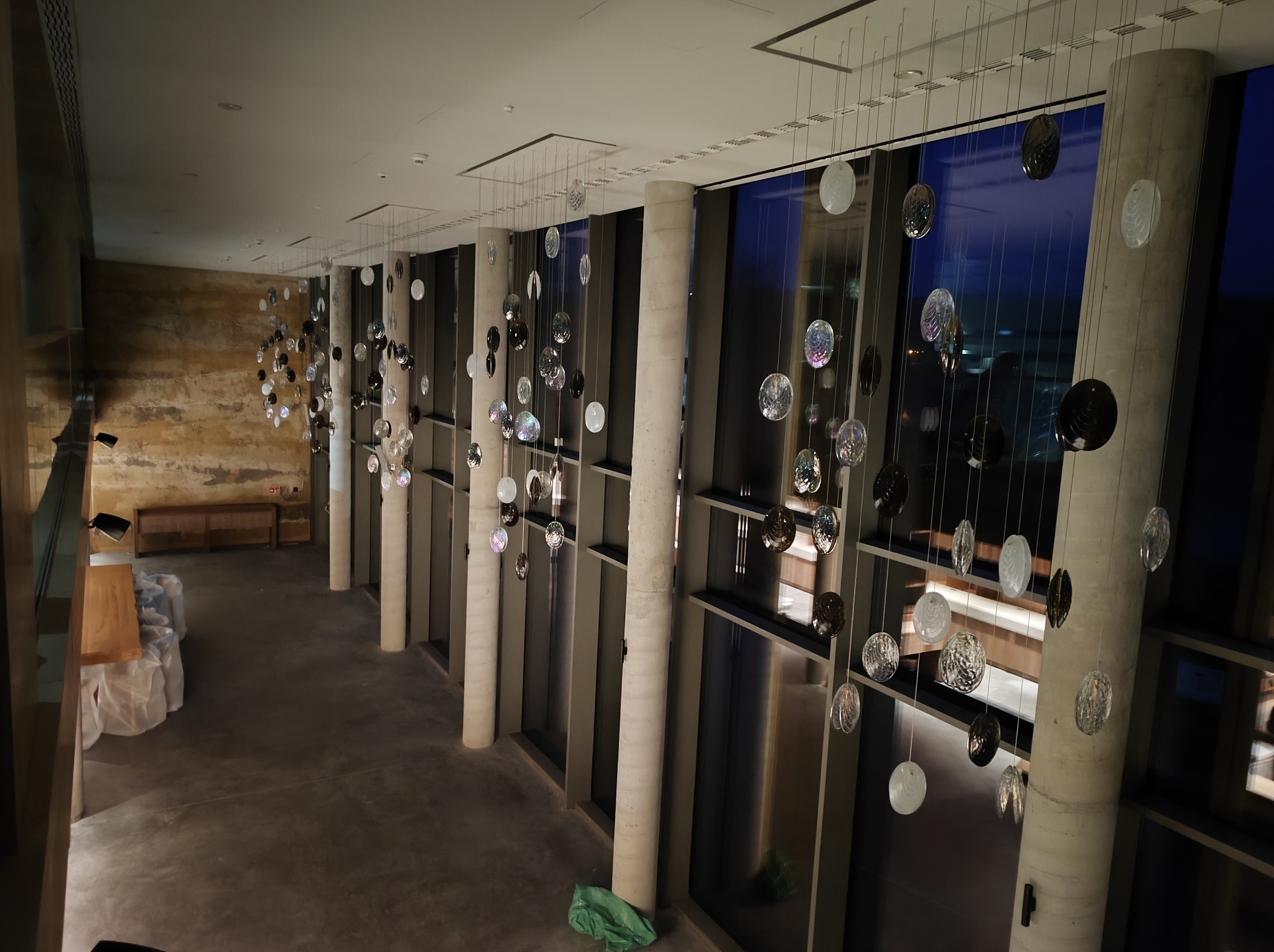 Wysoki hol z rzędami kolumn i oknami, ozdobiony wiszącymi dekoracjami świetlnymi w kształcie dysków, niektóre z nich z fakturą, zawieszone na cienkich linkach