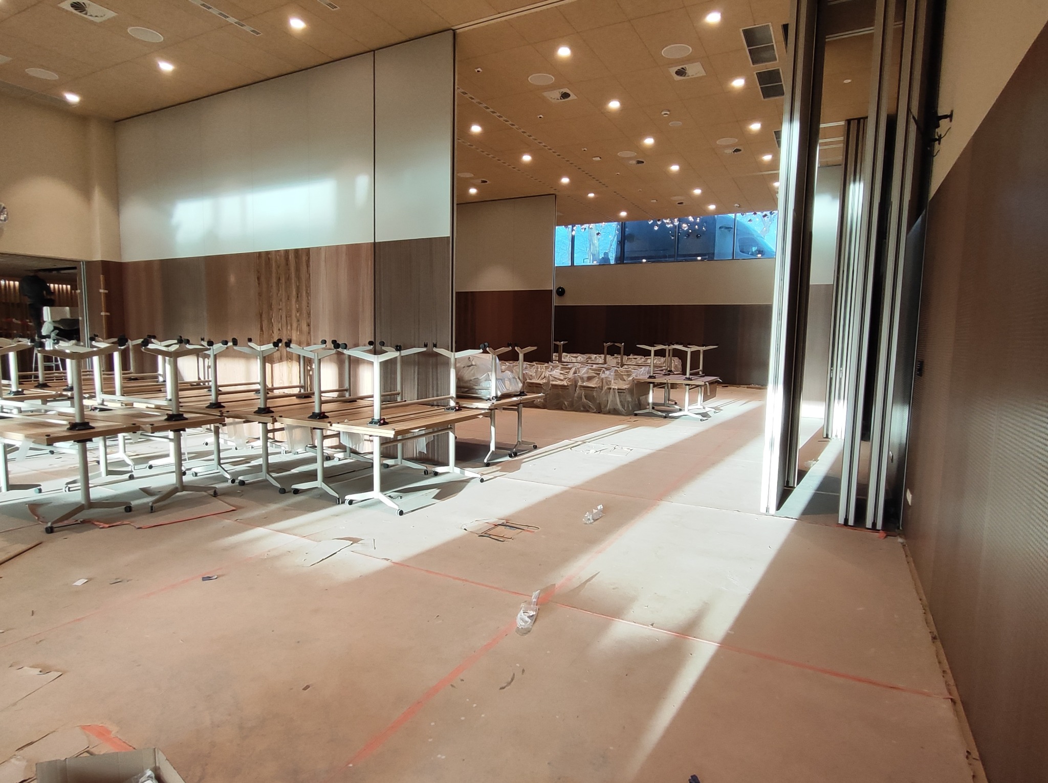 Przestronne wnętrze sali konferencyjnej w trakcie przygotowań, widoczne stoły z białymi nogami i drewnianymi blatami, rozłożone po całej powierzchni, z widocznymi elementami instalacji pod sufitem