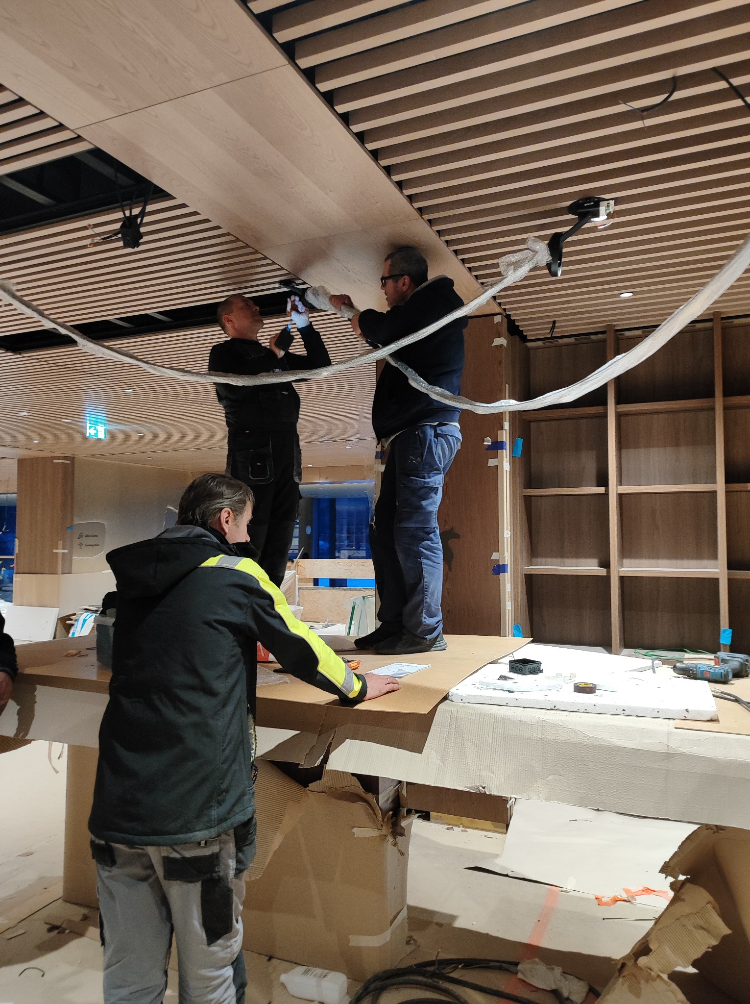 Dwóch monterów na podwyższeniu instaluje drewniane panele sufitowe, trzeci pracownik w odblaskowej kamizelce pomaga z dołu, widoczne narzędzia pracy i przewody oświetleniowe.