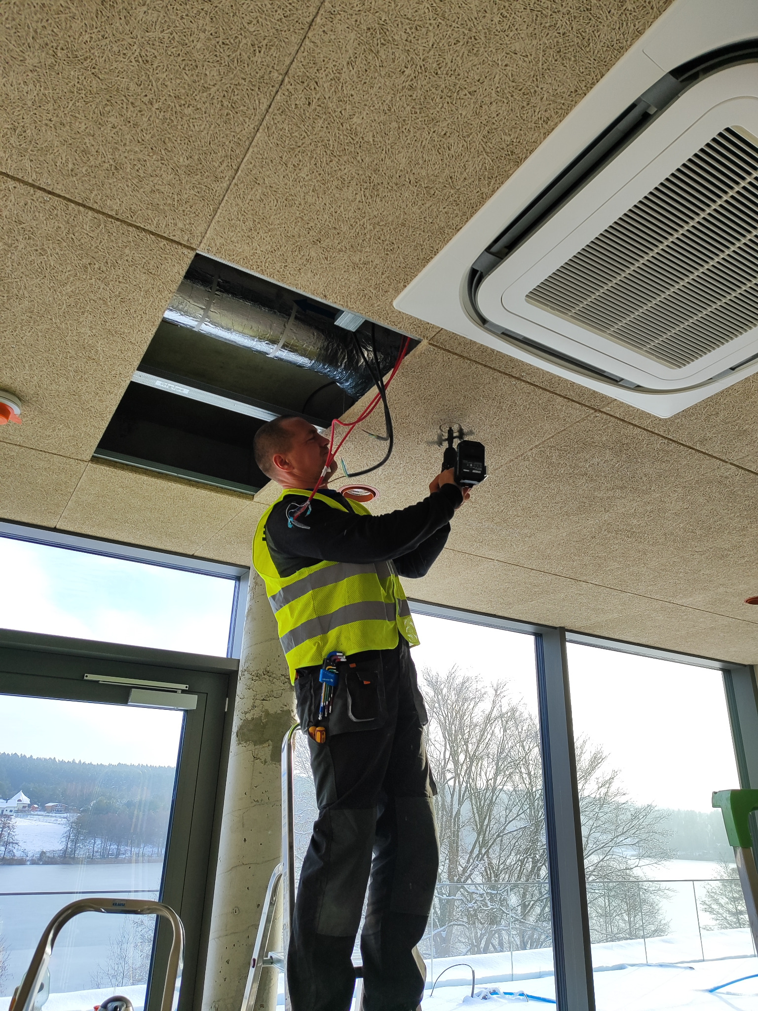 Elektryk w kamizelce odblaskowej na drabinie montuje oświetlenie sufitowe w nowoczesnym wnętrzu z widokiem na zimowy krajobraz.