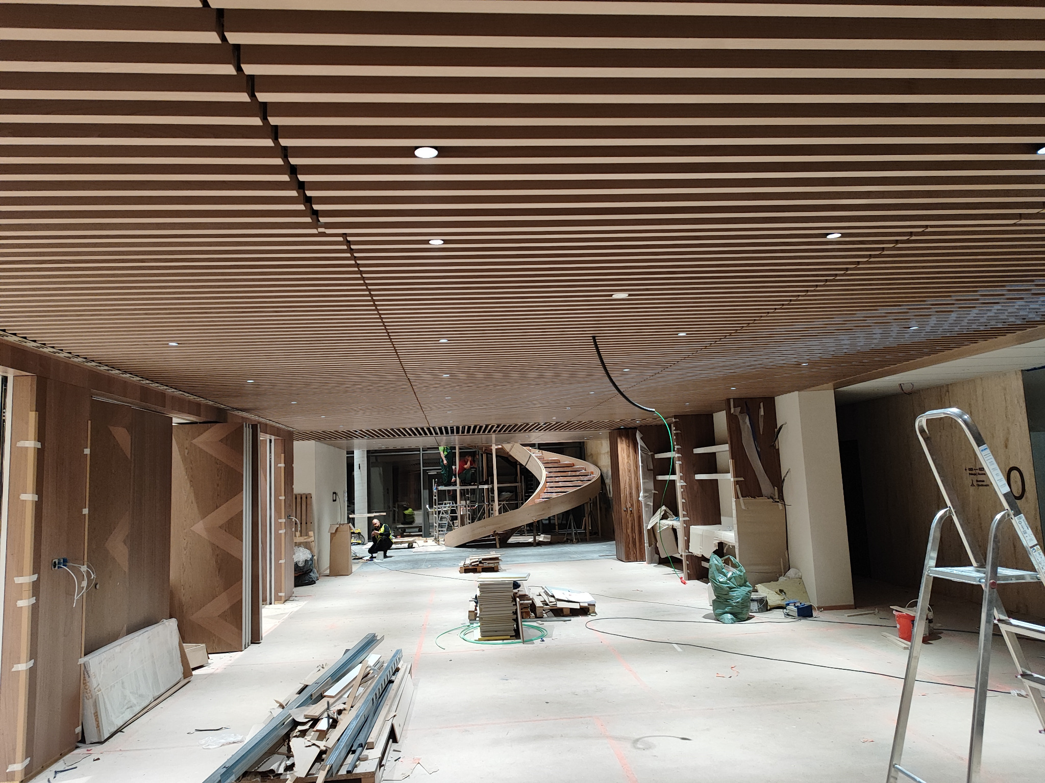 Wnętrze w trakcie wykańczania: drewniany sufit lamelowy z punktowymi lampami, spiralne schody w tle, widoczne elementy instalacji elektrycznej i drabina.
