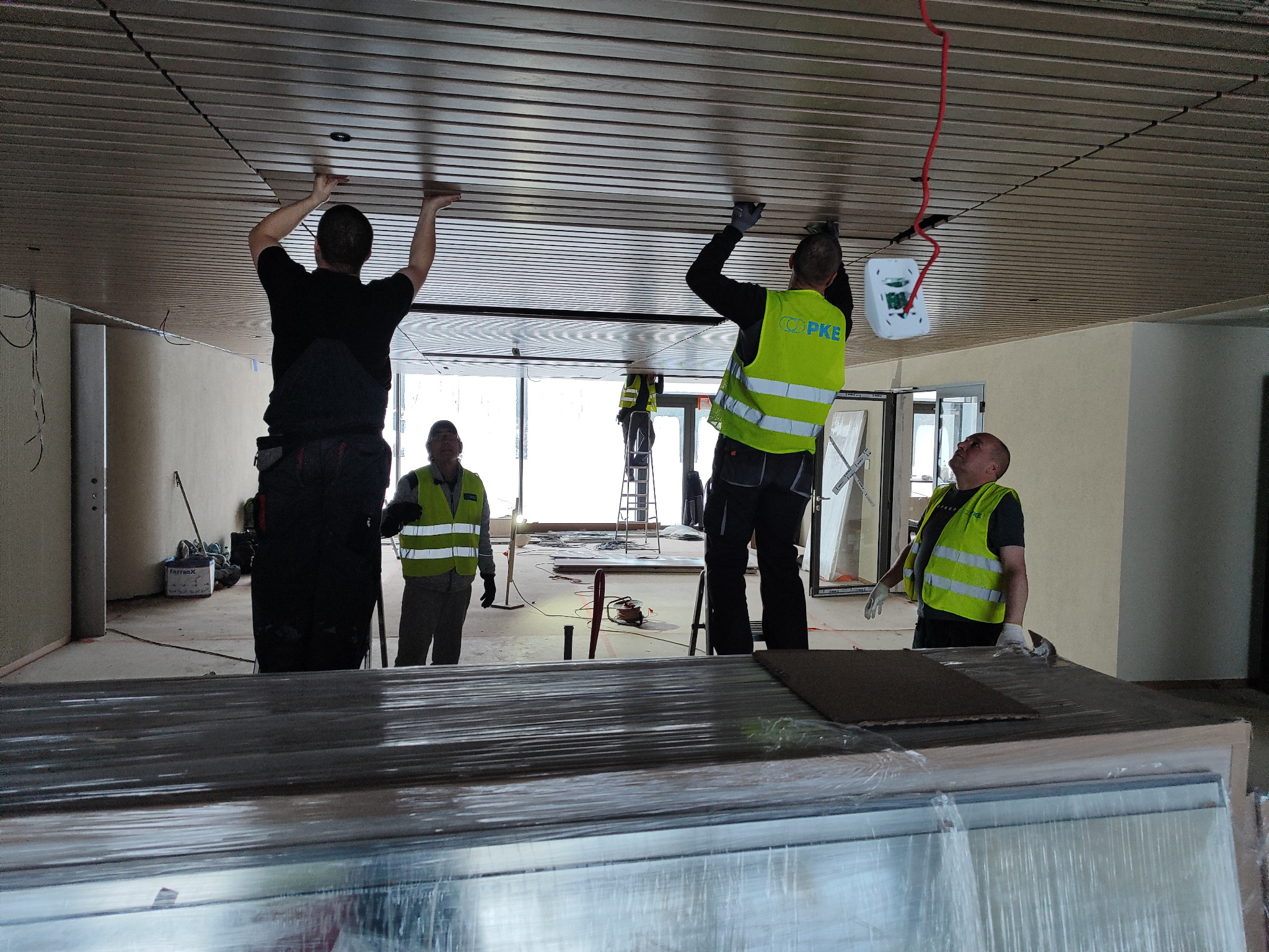 Pracownicy w odblaskowych kamizelkach montują drewniane panele sufitowe w nowoczesnym wnętrzu, widoczne przewody elektryczne i biała puszka instalacyjna.