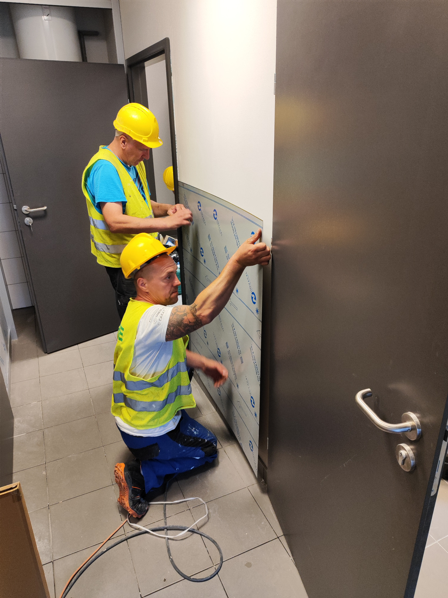 Dwóch monterów w żółtych kaskach i kamizelkach odblaskowych instaluje panel ścienny w pomieszczeniu z szarą podłogą i drzwiami.