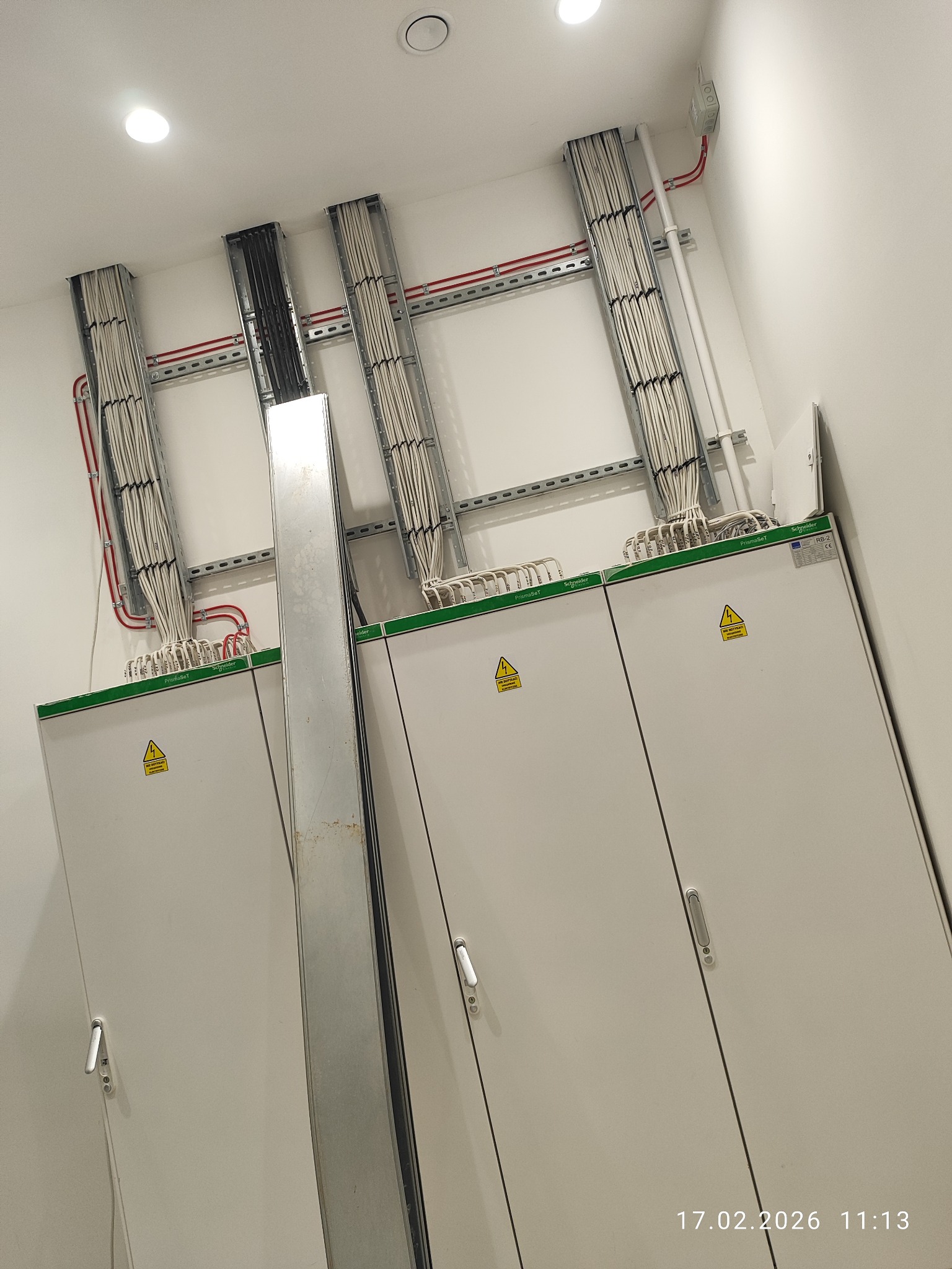 Rozdzielnice elektryczne z przewodami ułożonymi w korytkach, widoczne żółte znaki ostrzegawcze. Instalacja w nowym budynku.