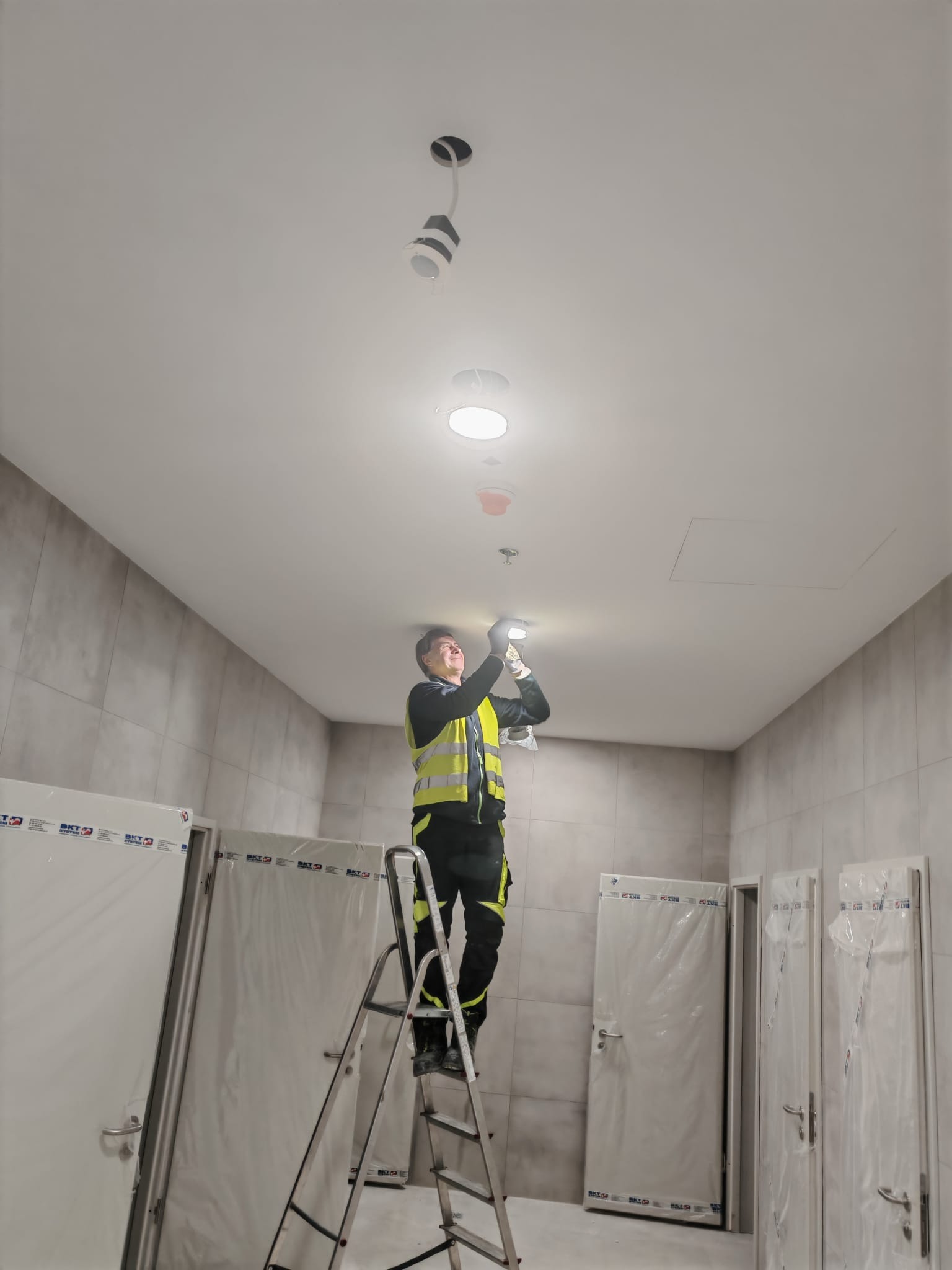 Elektryk na drabinie montuje oświetlenie sufitowe w pomieszczeniu z wykończonymi ścianami i zabezpieczonymi folią drzwiami. Widoczne oprawy i przewody.