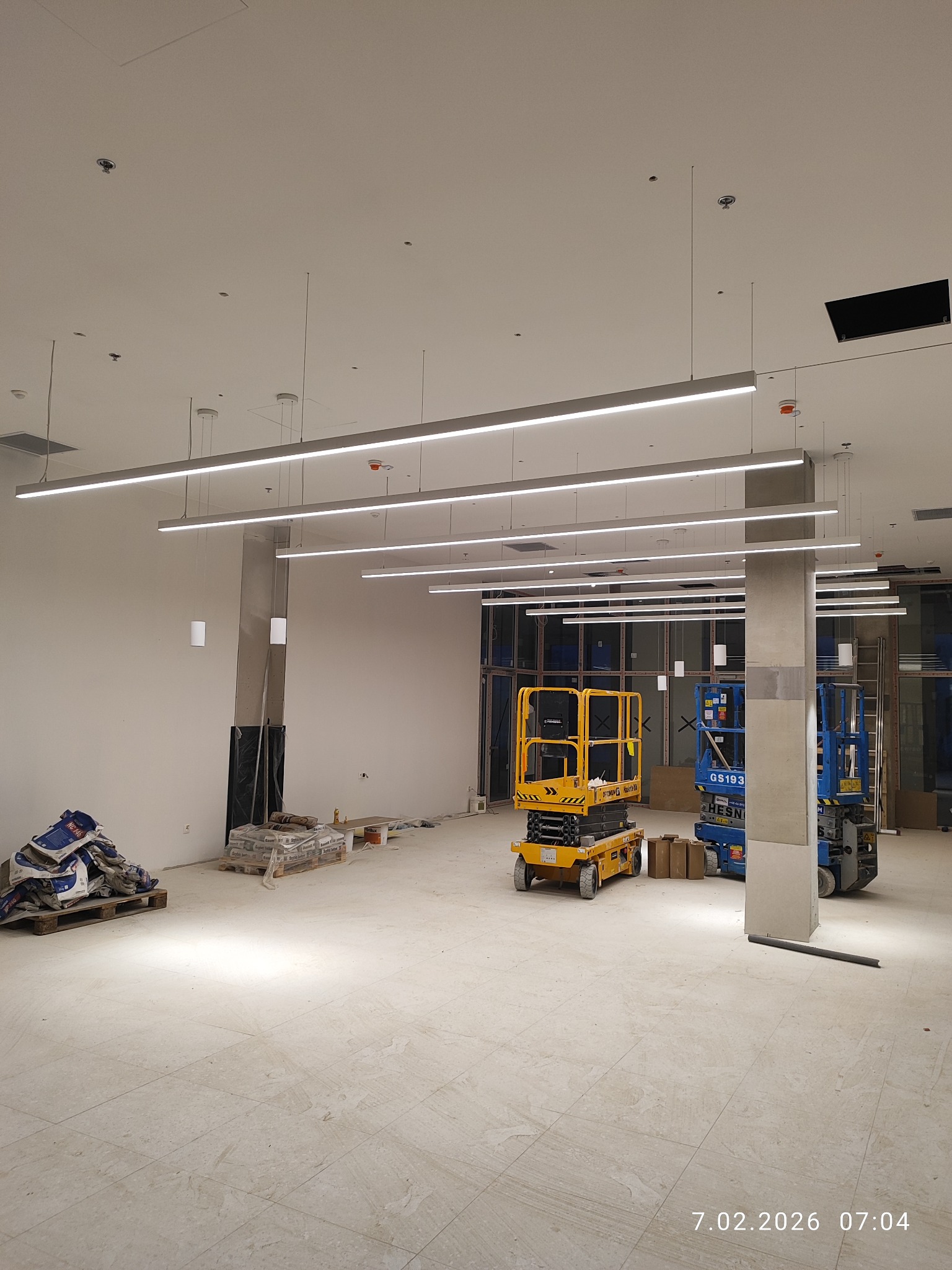 Montaż podłużnych lamp LED w nowym lokalu. Dwie podnośniki koszowe, żółty i niebieski, stoją na posadzce. Worki z materiałami budowlanymi przy ścianie.