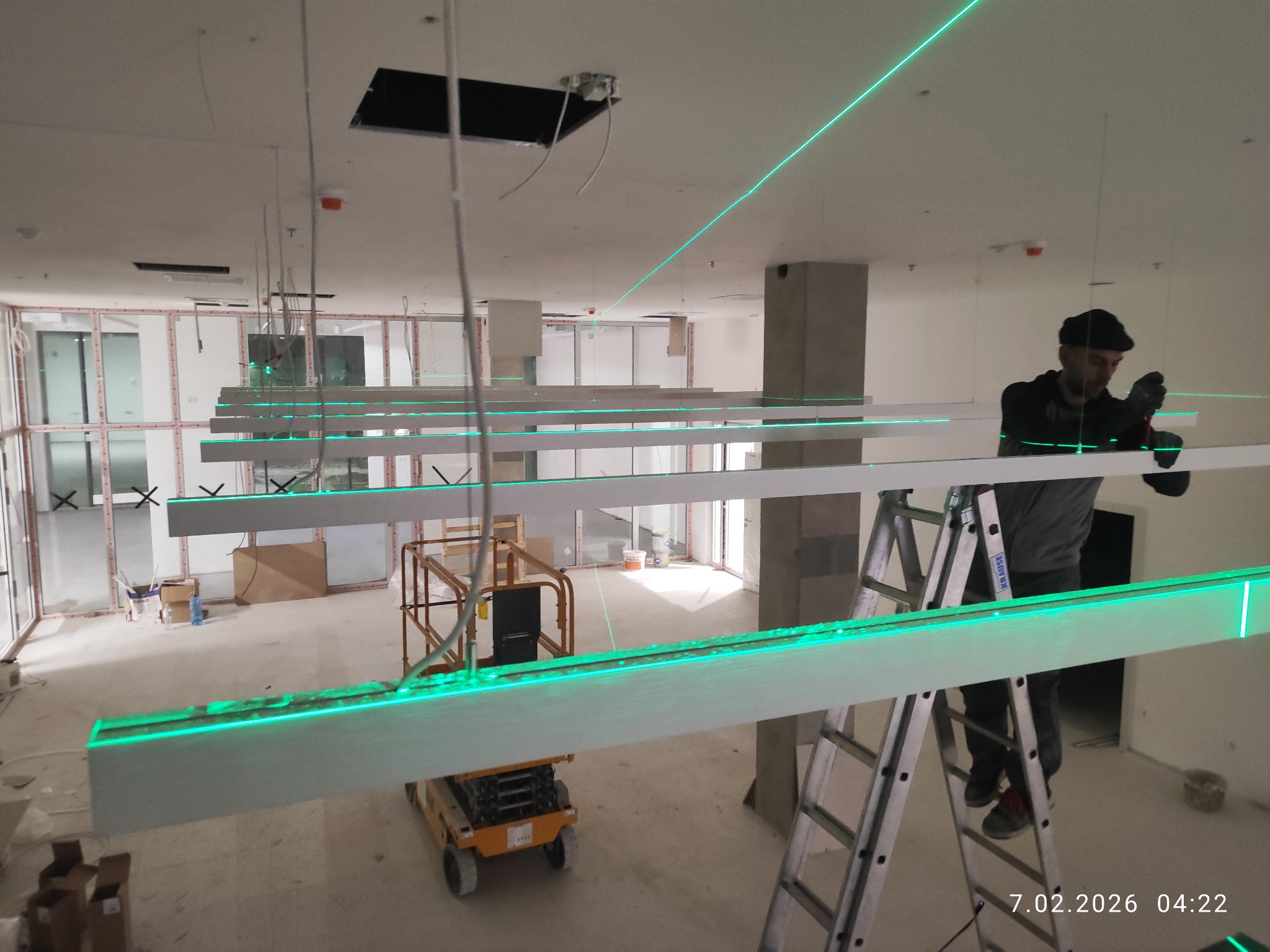 Montera na drabinie poziomuje laserowo podwieszane, długie panele oświetleniowe LED w surowym wnętrzu biurowym. Widoczny podnośnik i zielone linie lasera.