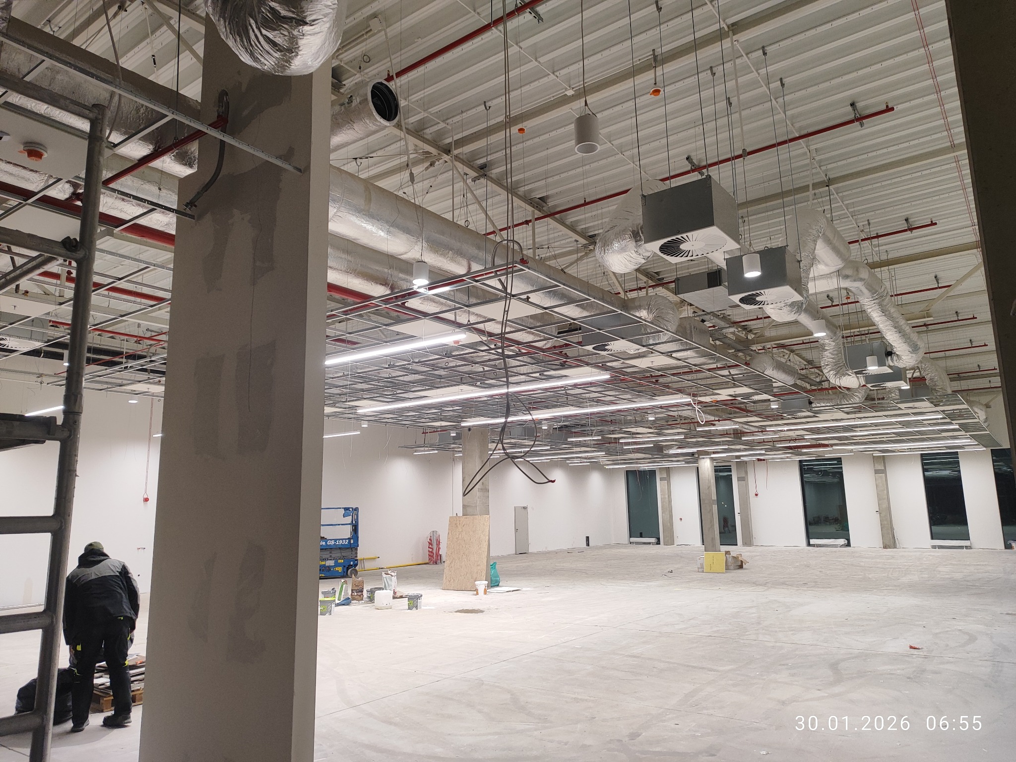 Wnętrze hali w trakcie prac wykończeniowych. Widoczne instalacje pod sufitem, kolumna w szpachli, drabina i pracownik w oddali. Oświetlenie ledowe zamontowane.
