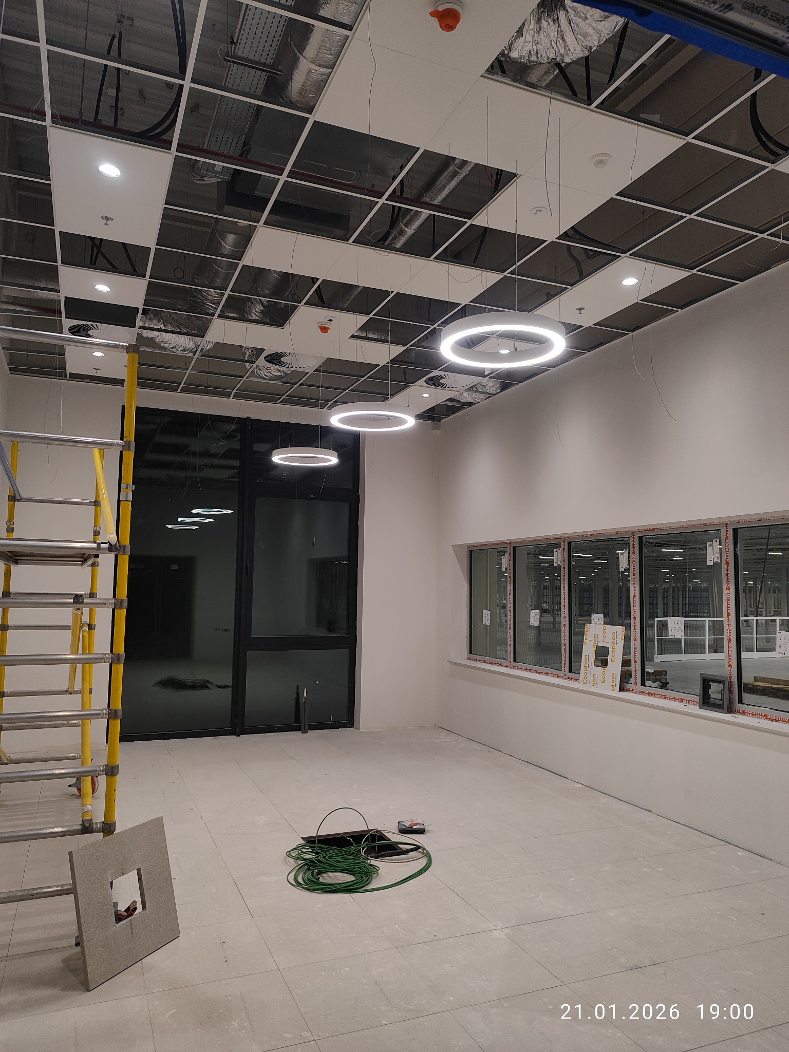 Wnętrze w trakcie montażu oświetlenia: podwieszany sufit z rastrem, okrągłe lampy LED, przewody, rusztowanie. Widok na okna i kable na podłodze.