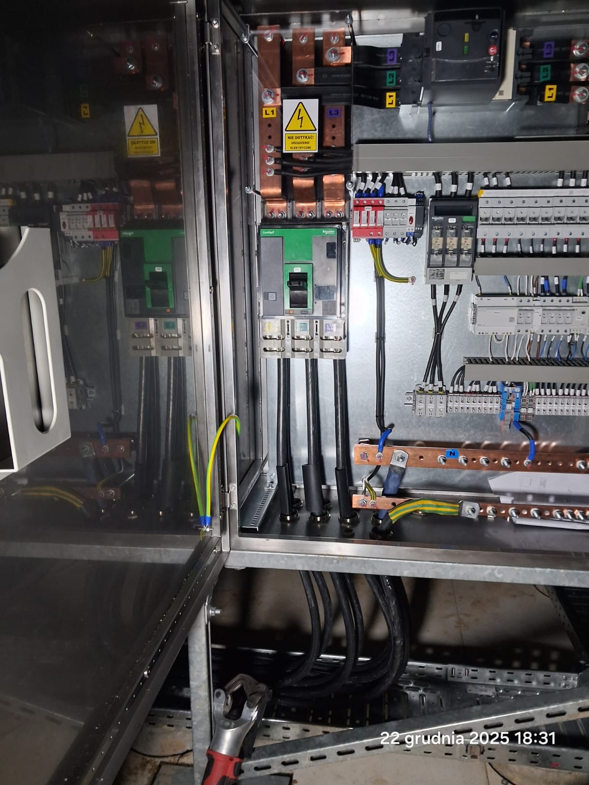 Wnętrze szafy elektrycznej z zainstalowanymi bezpiecznikami, przewodami i szynami. Widoczne oznaczenia ostrzegawcze i narzędzia. Metalowa konstrukcja, precyzyjne połączenia.