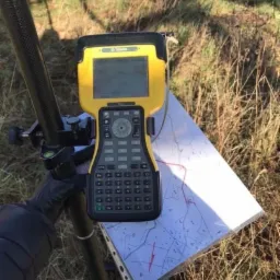 Żółty odbiornik GPS Trimble na statywie, z mapą w tle, podczas pomiarów w terenie.