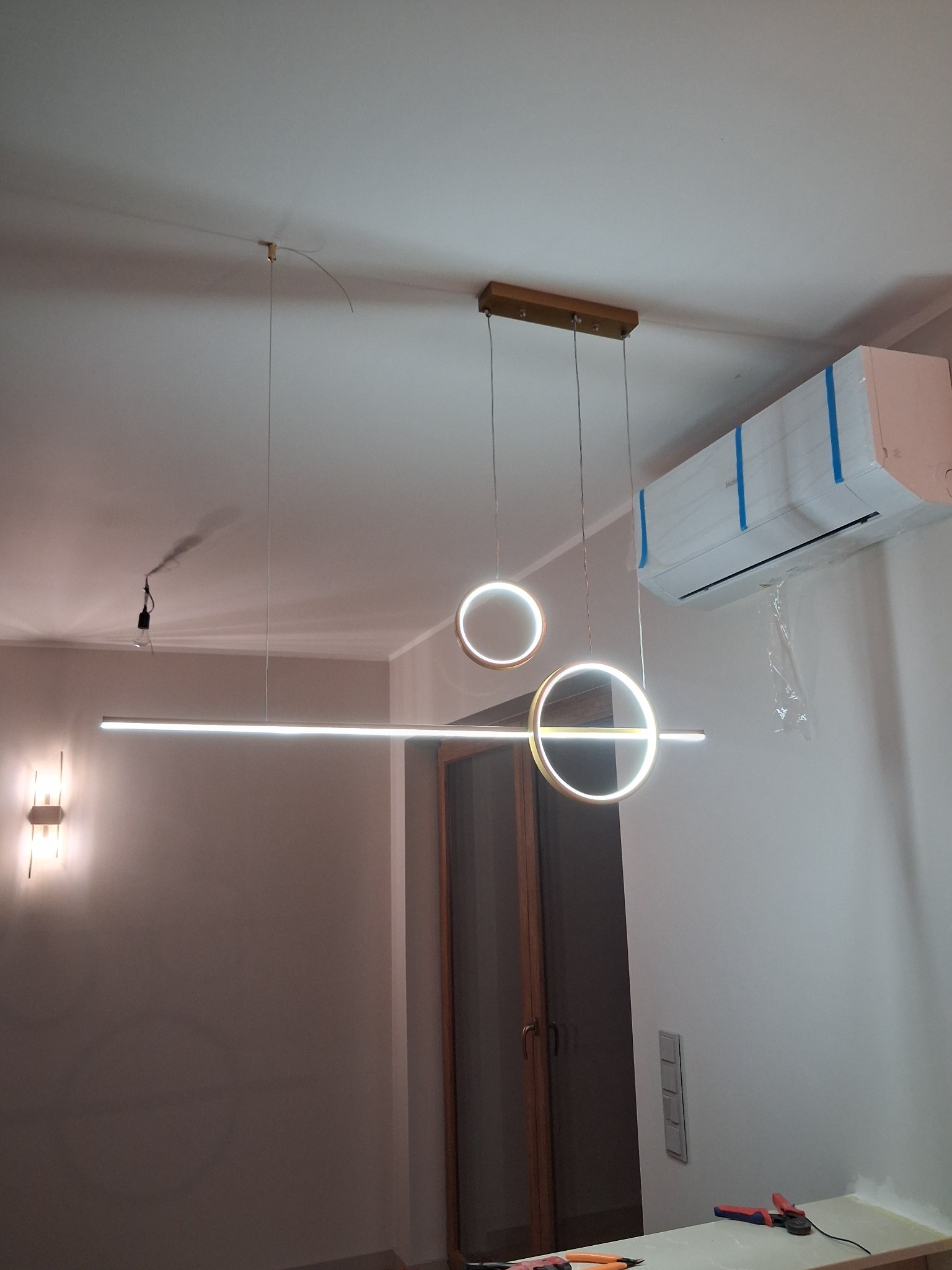 Nowoczesna lampa wisząca LED z okrągłymi elementami, zamontowana w pomieszczeniu z klimatyzacją. Widoczne narzędzia na blacie sugerują prace instalacyjne.