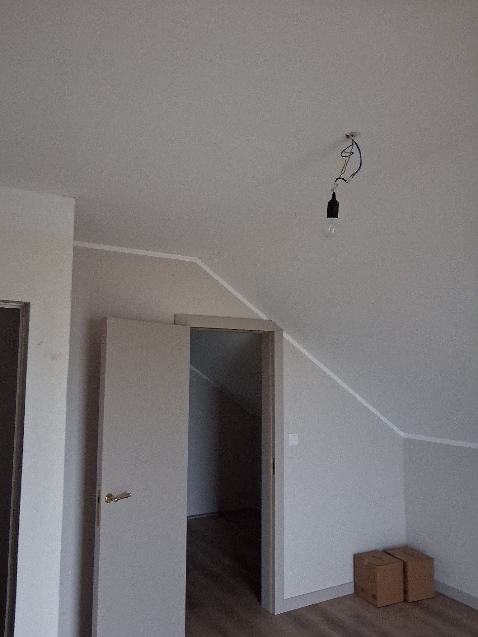 Poddasze po tynkowaniu: szare ściany, białe listwy, surowa żarówka, drzwi wewnętrzne i dwa kartony na podłodze. Widok na wykończone wnętrze.