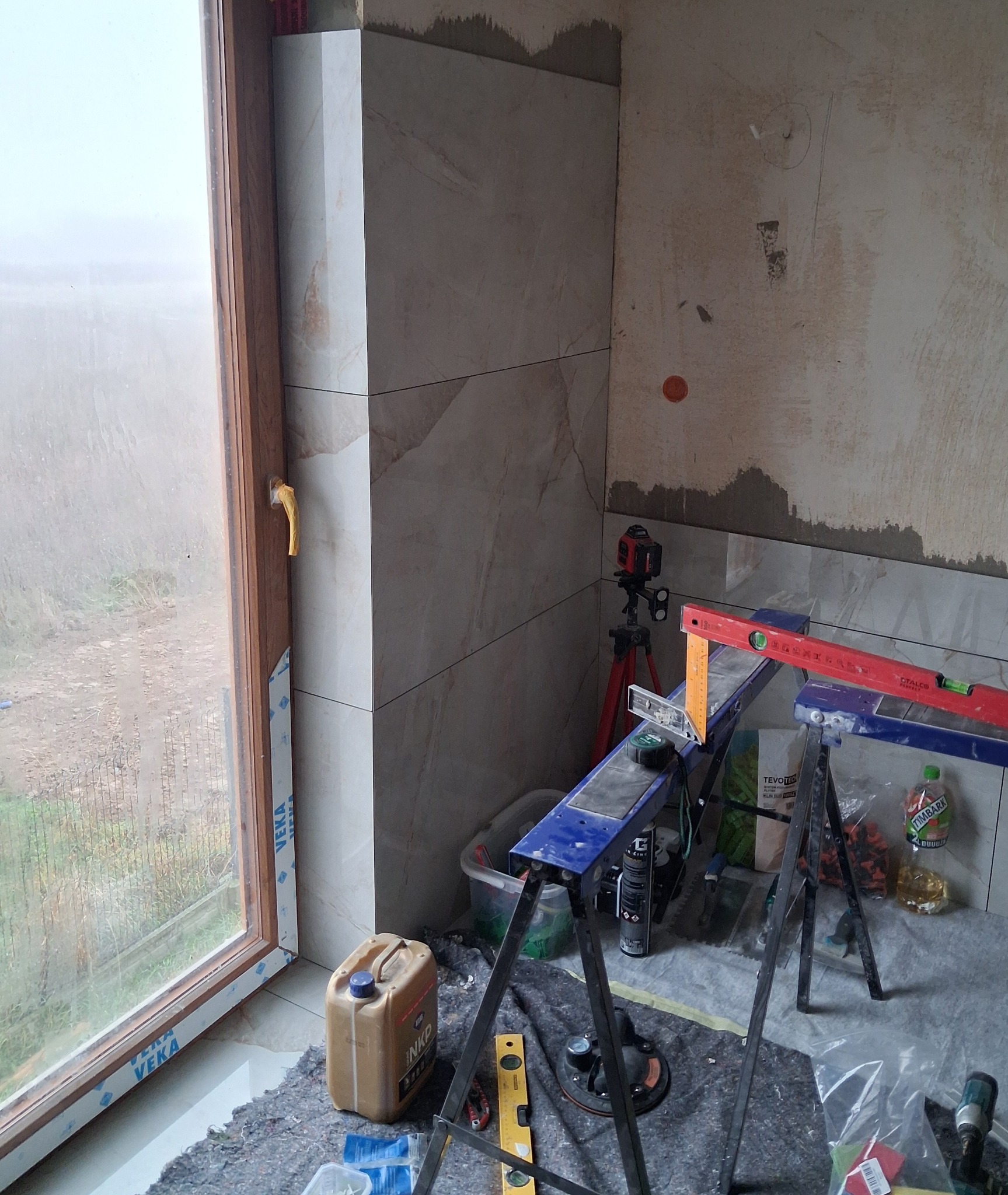 Wykończenie narożnika płytkami imitującymi marmur, widoczne narzędzia glazurnicze i poziomica laserowa. Prace wykończeniowe w pomieszczeniu z oknem.