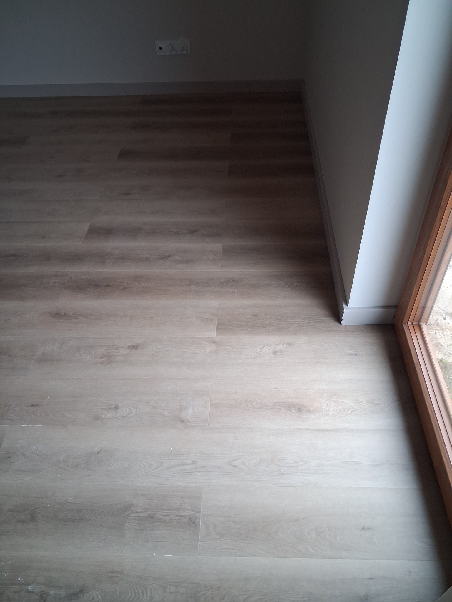 Jasne panele podłogowe ułożone w rogu pokoju, widoczne listwy przypodłogowe i fragment okna z drewnianą ramą. Świeżo wykończone wnętrze.