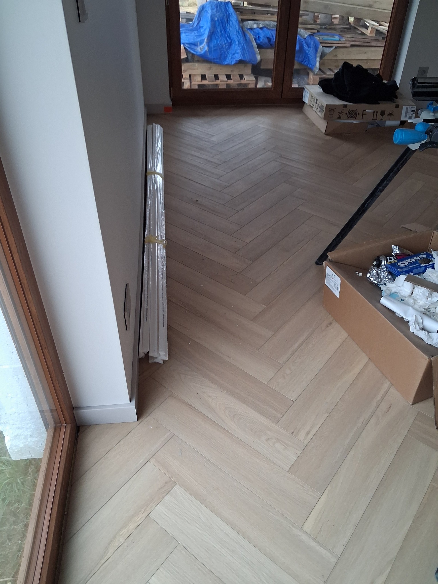 Jasna podłoga ułożona w jodełkę francuską, widok z góry. Przy ścianie listwy przypodłogowe i okno. W tle okno balkonowe i materiały budowlane.