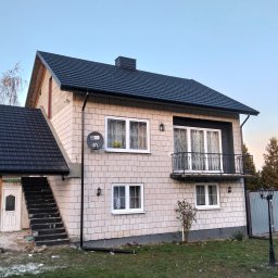 Dwukondygnacyjny dom z jasnej cegły z ciemnym dachem dwuspadowym i balkonem. Widoczne schody zewnętrzne z zadaszeniem i lampa uliczna. Tło: drzewa i niebo.