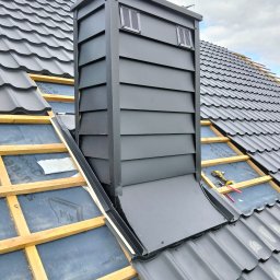 Montaż czarnego komina wentylacyjnego na dachu z czarnej blachodachówki. Widoczne drewniane łaty i membrana dachowa. Częściowo pokryty dach.