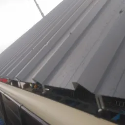 Szare, trapezowe blachy dachowe z widocznym systemem rynnowym, zamontowane na hali przemysłowej, częściowo mokre od deszczu, z widocznym żurawiem w tle.