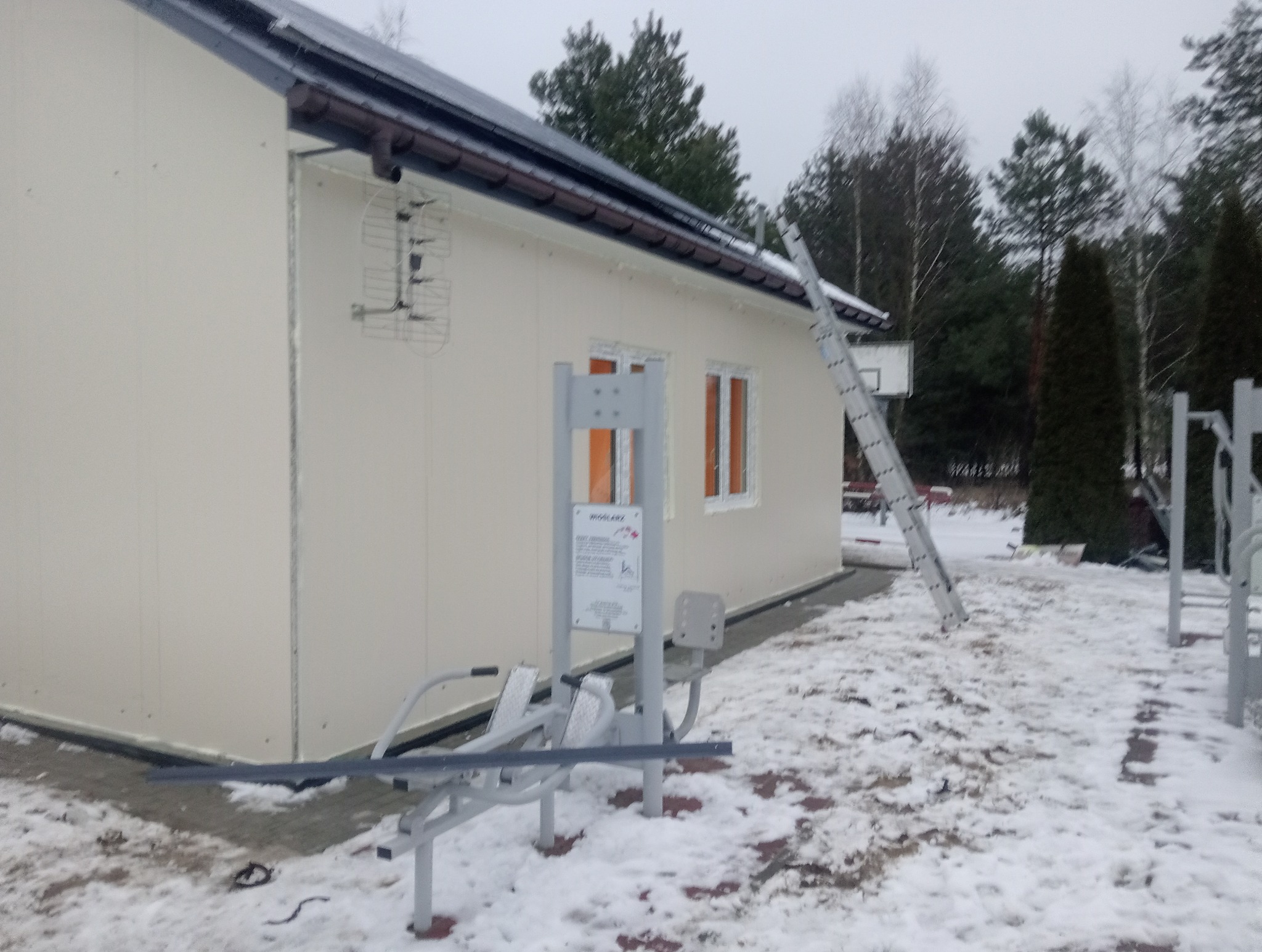 Budynek z płyt warstwowych z anteną na ścianie, drabina oparta o ścianę, obok urządzenia do ćwiczeń na zewnątrz, zimowy krajobraz.