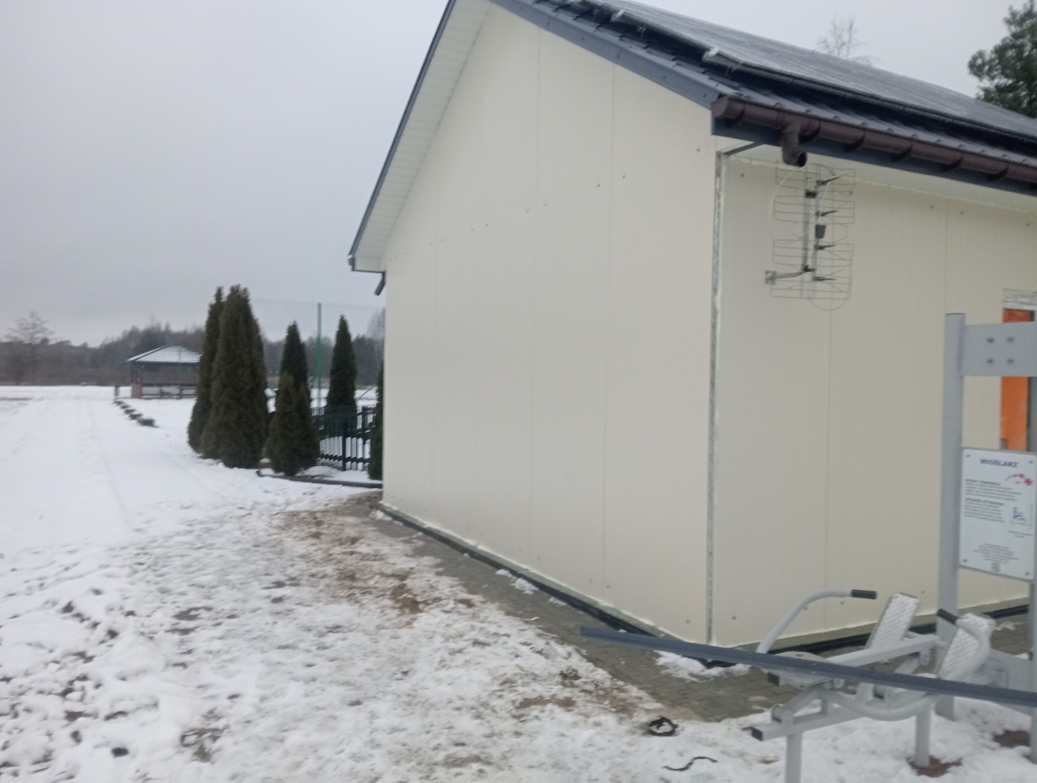 Boczny widok hali z płyt warstwowych w zimowej scenerii, widoczna antena na ścianie i sprzęt do ćwiczeń na zewnątrz.