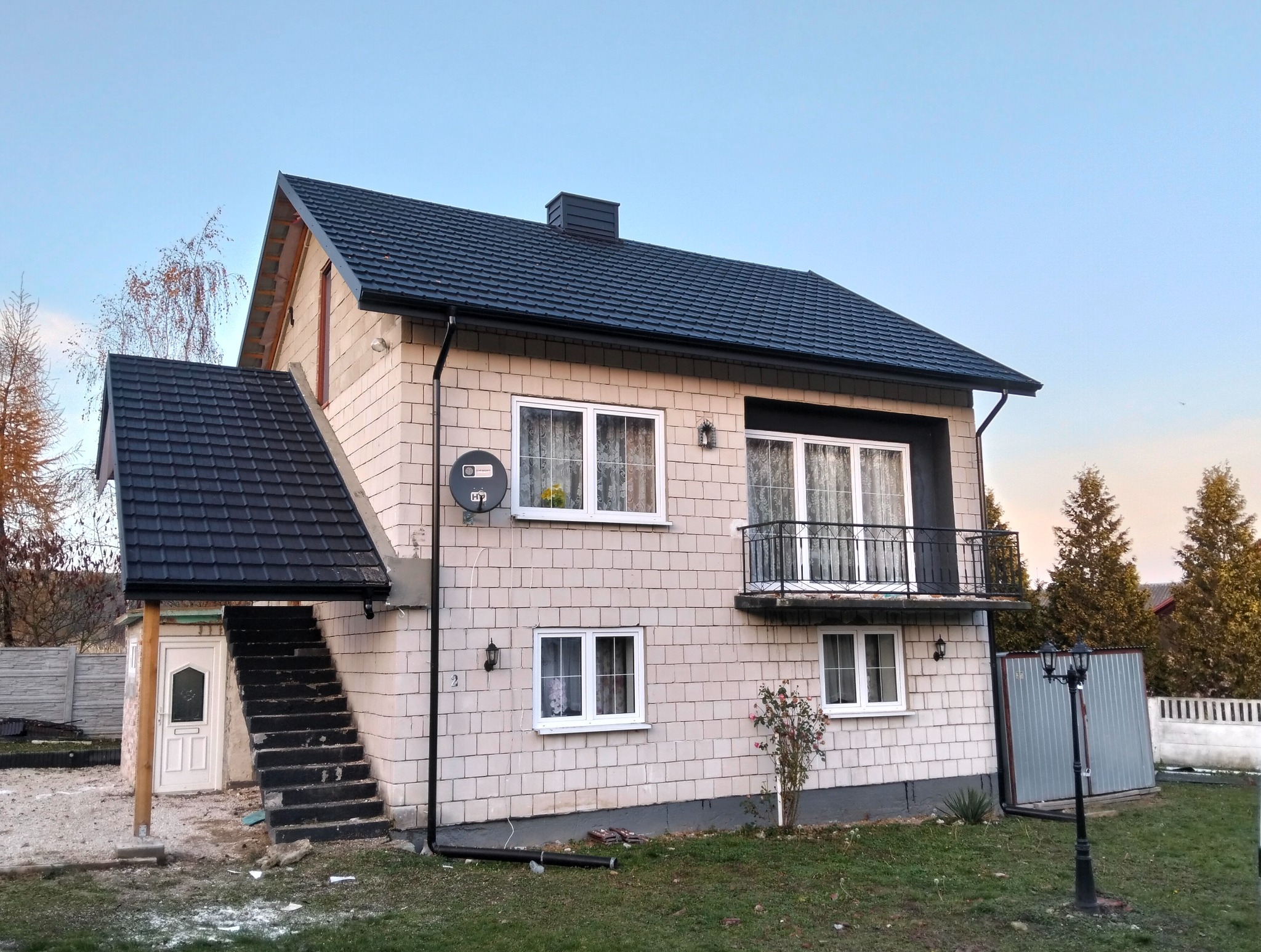 Dwukondygnacyjny dom z jasnej cegły z ciemnym dachem dwuspadowym i balkonem. Widoczne schody zewnętrzne z zadaszeniem i lampa uliczna. Tło: drzewa i niebo.