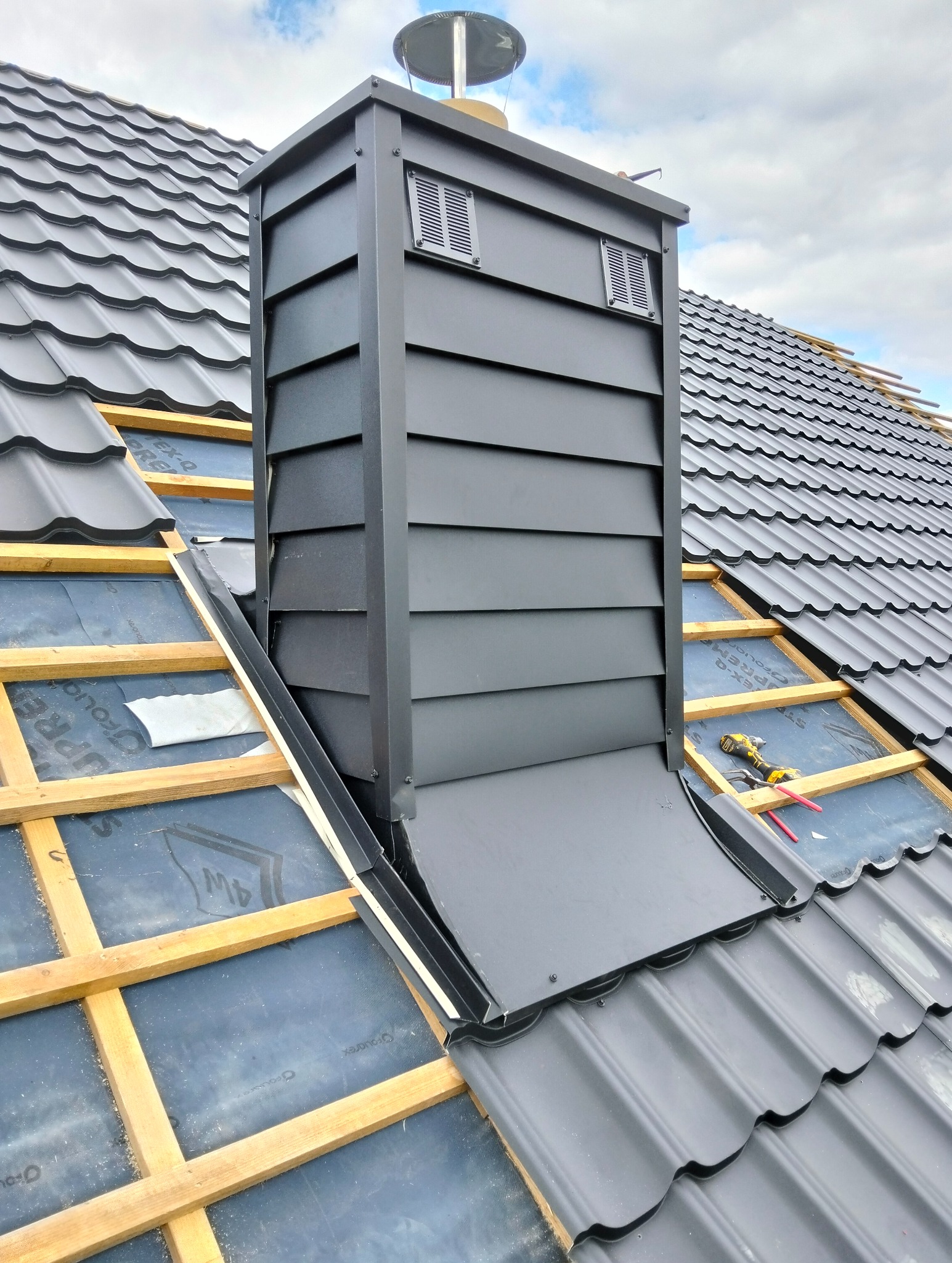 Montaż czarnego komina wentylacyjnego na dachu z czarnej blachodachówki. Widoczne drewniane łaty i membrana dachowa. Częściowo pokryty dach.