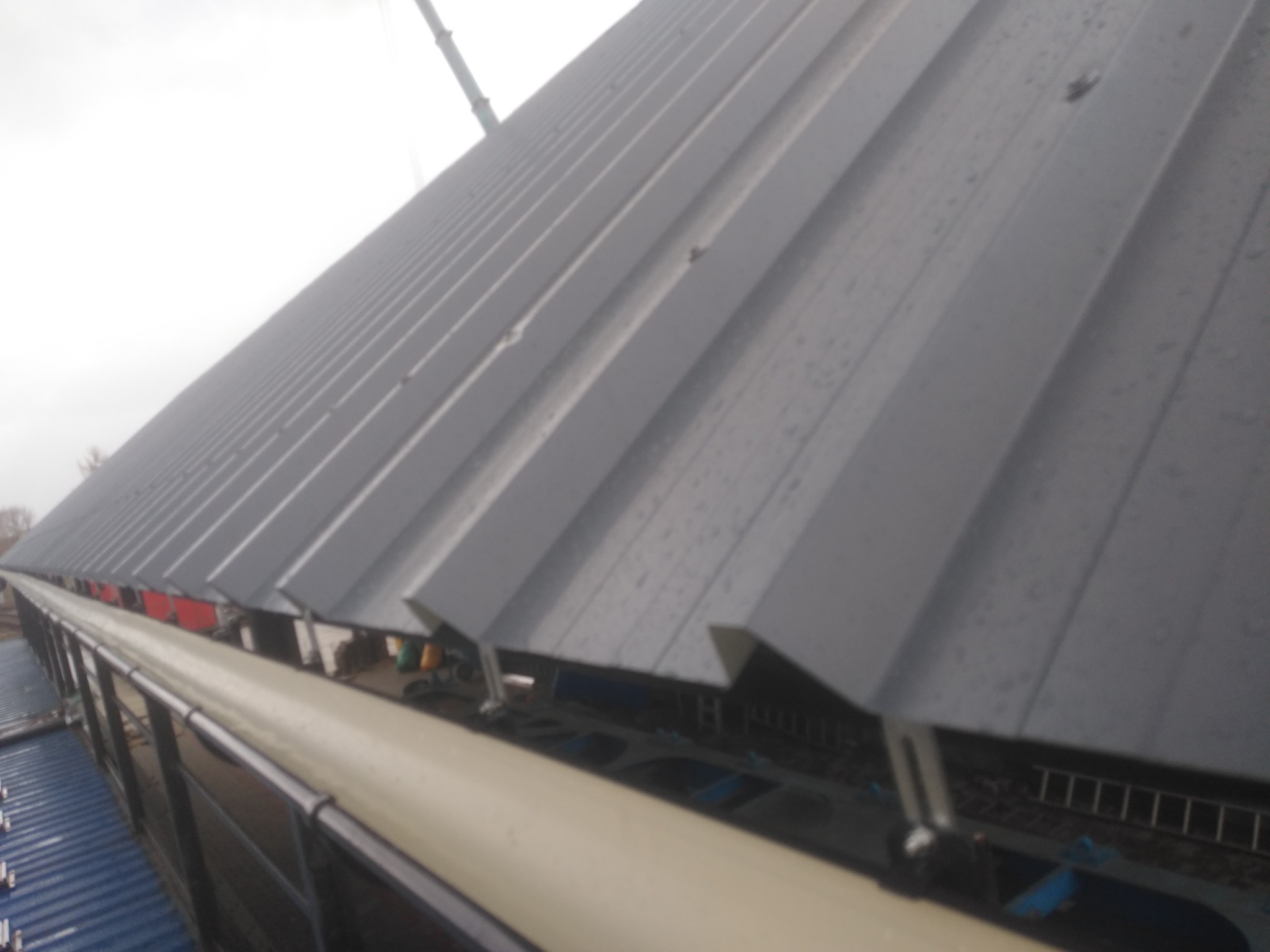 Szare, trapezowe blachy dachowe z widocznym systemem rynnowym, zamontowane na hali przemysłowej, częściowo mokre od deszczu, z widocznym żurawiem w tle.