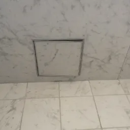 Biała marmurowa ściana i podłoga z ukrytym, kwadratowym włazem rewizyjnym z metalową ramką.