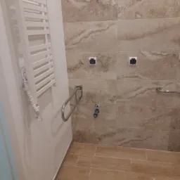 Niedokończona łazienka z beżowymi płytkami imitującymi marmur, zamontowanym grzejnikiem drabinkowym, uchwytem dla niepełnosprawnych, gniazdkami elektrycznymi i wyprowadzonymi przyłączami...
