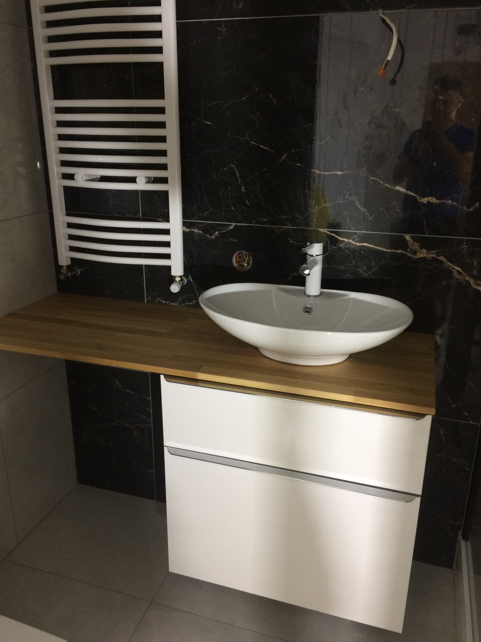 Nowoczesna łazienka z umywalką nablatową na drewnianym blacie, białą szafką pod umywalką, grzejnikiem drabinkowym i czarnymi płytkami imitującymi marmur z widocznymi przewodami elektrycznymi.
