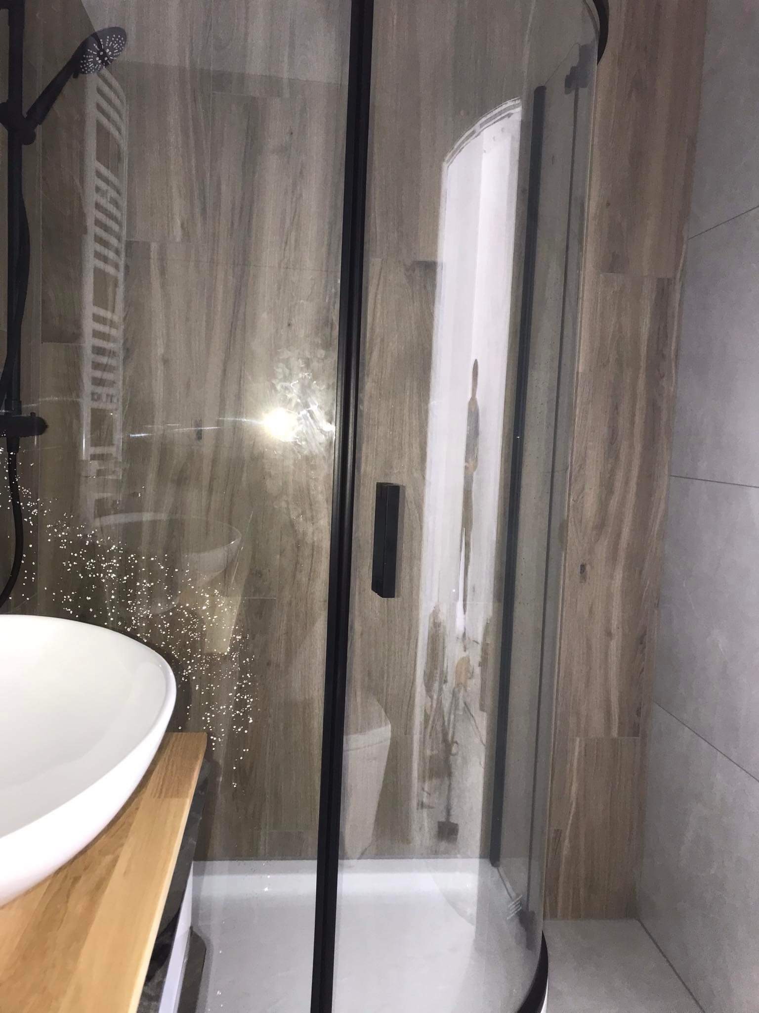 Nowoczesna łazienka z kabiną prysznicową z czarnymi ramami, umywalką nablatową na drewnianym blacie i płytkami imitującymi drewno na ścianie.