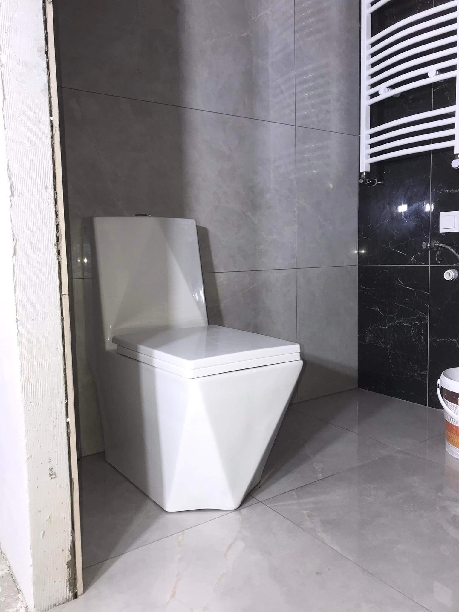 Nowoczesna toaleta wisząca o geometrycznym kształcie w łazience z szarymi i czarnymi płytkami imitującymi marmur, widoczny fragment niedokończonego ościeża drzwi.