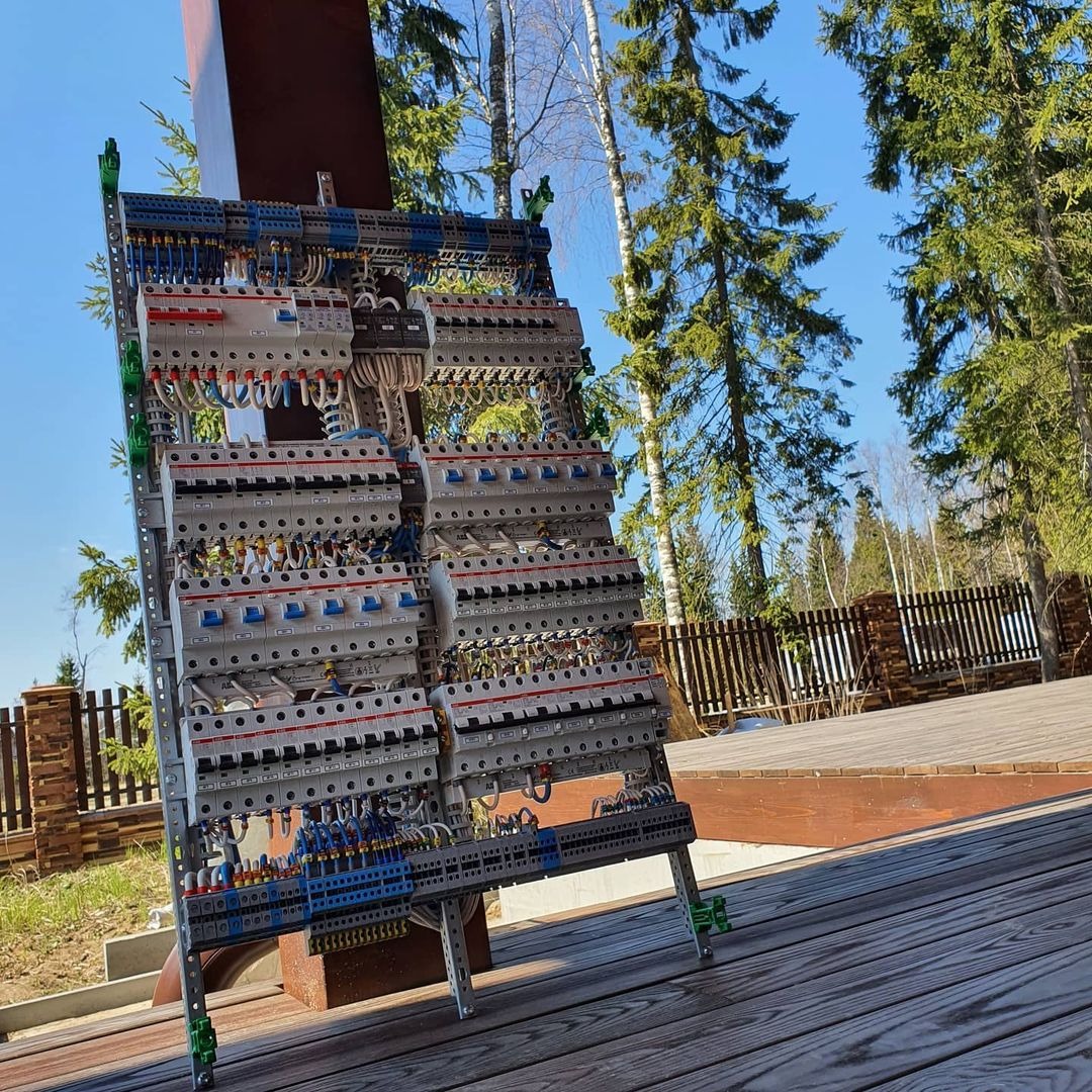 Rozdzielnica elektryczna na metalowym stojaku, widoczne liczne zabezpieczenia, przewody i złącza na tle drewnianego tarasu i zieleni.