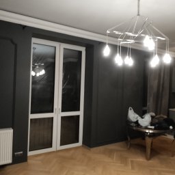 Kamil Kawiecki - Nowoczesne wnętrze z ciemnoszarymi ścianami z dekoracyjnymi panelami, białymi listwami przysufitowymi i grzejnikiem, parkietem w jodełkę, przeszklonymi drzwiami balkonowymi i designerską lampą...