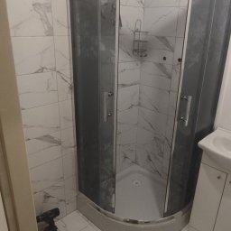 Kamil Kawiecki - Narożna kabina prysznicowa z szarymi szybami w kwiatowy wzór, wykończona marmurowymi płytkami, na podłodze narzędzia i silikon.