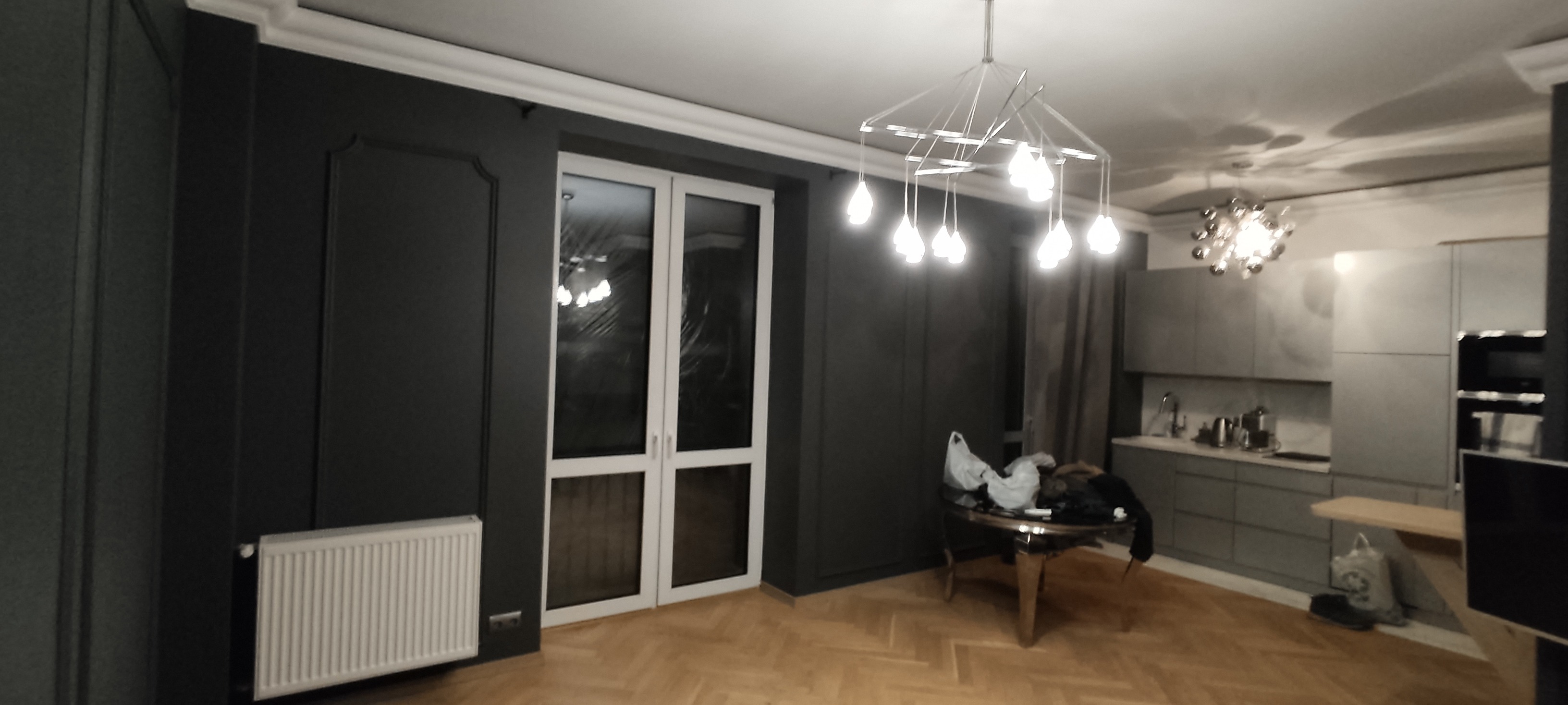 Nowoczesne wnętrze z ciemnoszarymi ścianami z dekoracyjnymi panelami, białymi listwami przysufitowymi i grzejnikiem, parkietem w jodełkę, przeszklonymi drzwiami balkonowymi i designerską lampą...