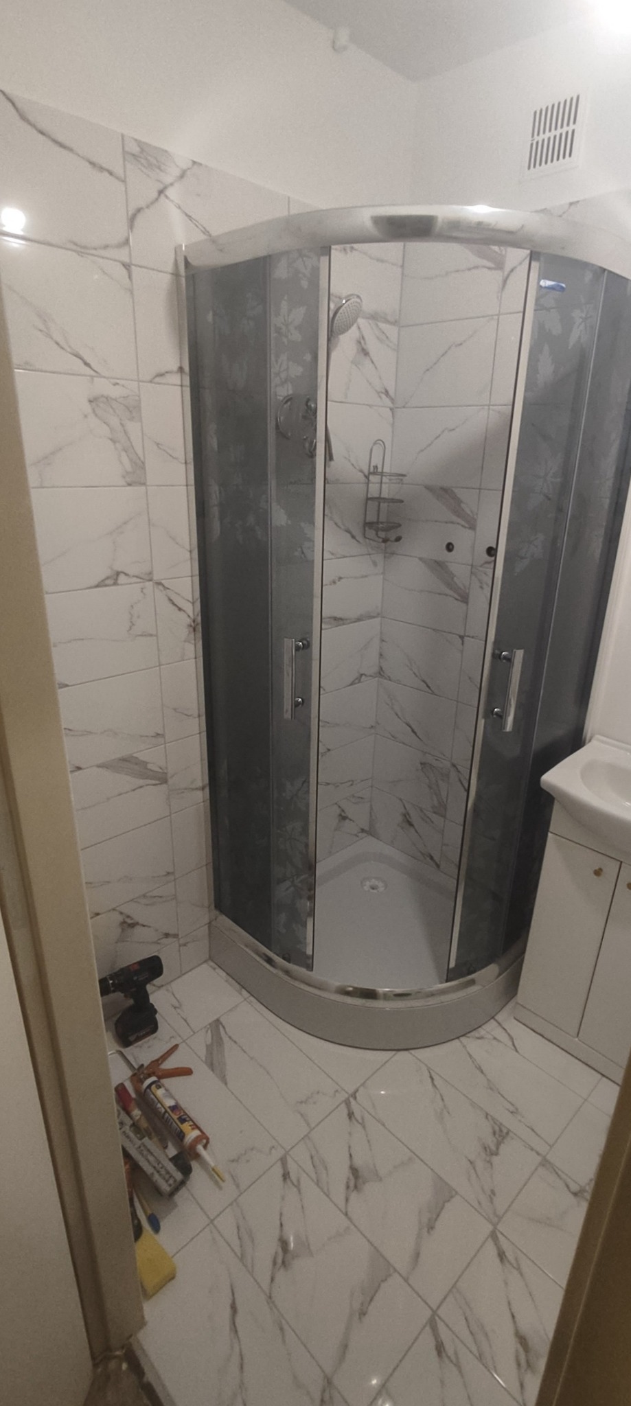 Narożna kabina prysznicowa z szarymi szybami w kwiatowy wzór, wykończona marmurowymi płytkami, na podłodze narzędzia i silikon.